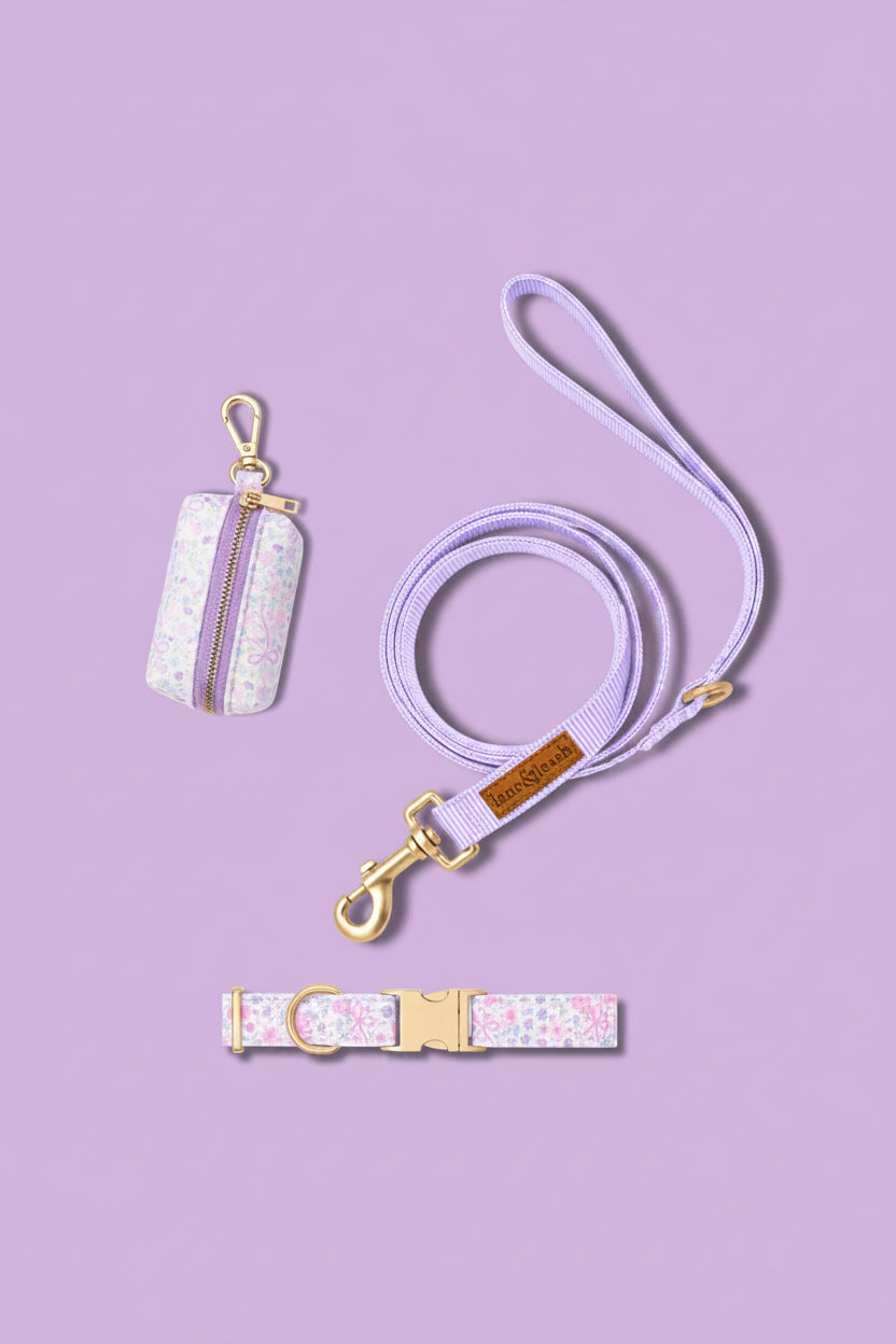 Ribbon Garden collection accessories flat lay on a lavender background: waste bag holder in pink floral print, matching purple leash, and pink floral print dog collar with gold hardware