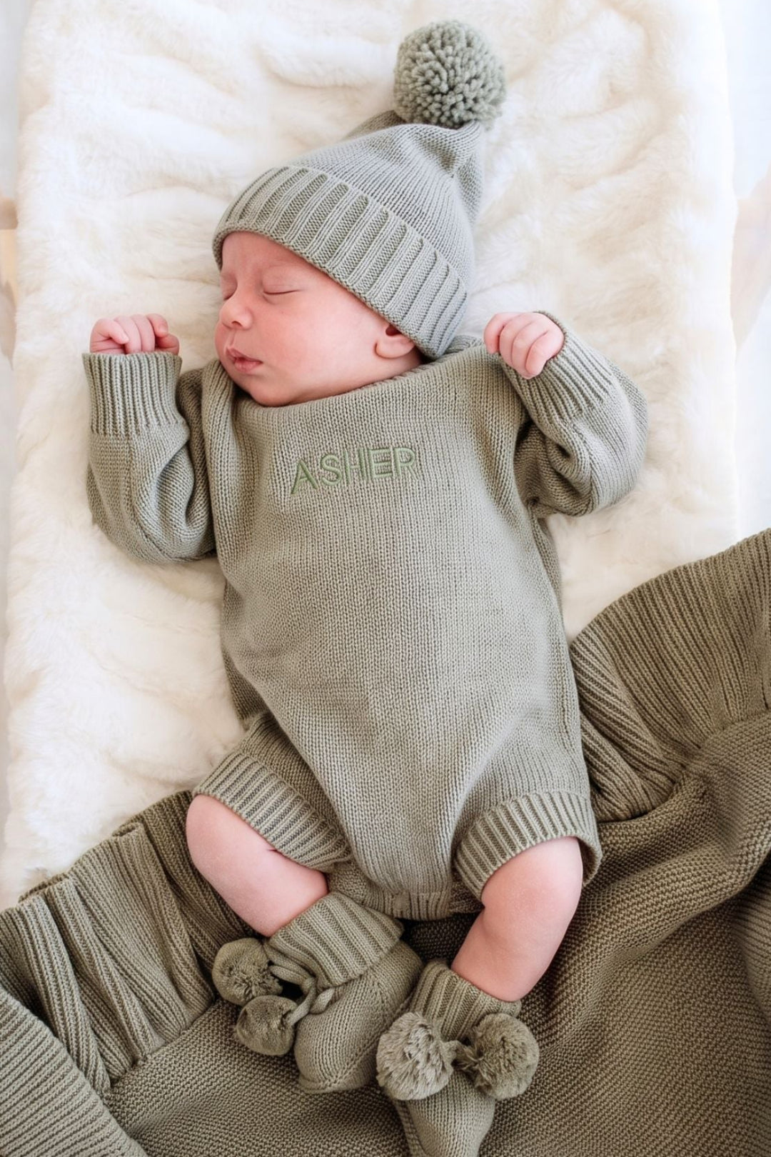 A sleeping newborn baby wearing a Caden Lane olive green knit onesie with 'ASHER' embroidered on the chest, a matching knit pom-pom hat, and booties, lying on a white blanket.