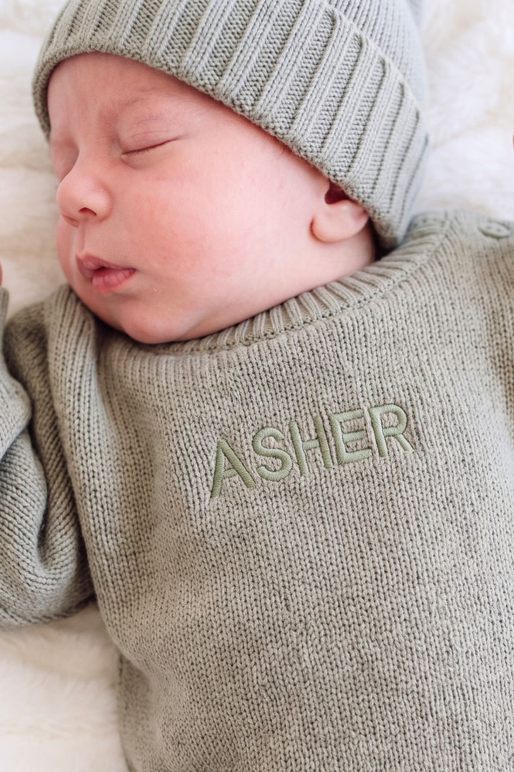 Close-up of sleeping baby wearing Caden Lane sage green knit romper with "Asher" name embroidery and matching ribbed knit hat