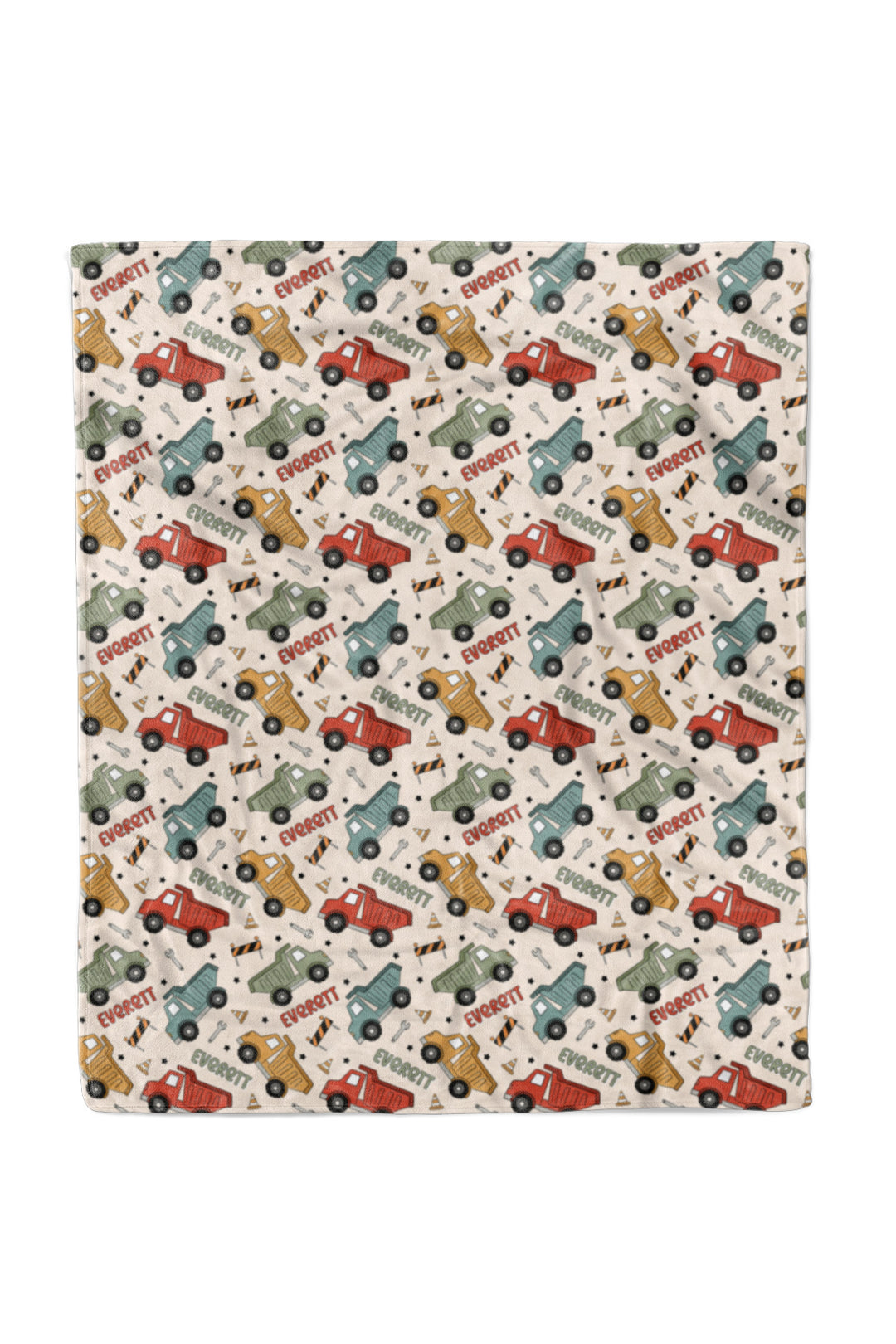 Flat lay of a Caden Lane cream plush blanket in a Busy Builder print featuring red, mustard, sage, and blue dump trucks, reading "Everett" in alternating red and green.