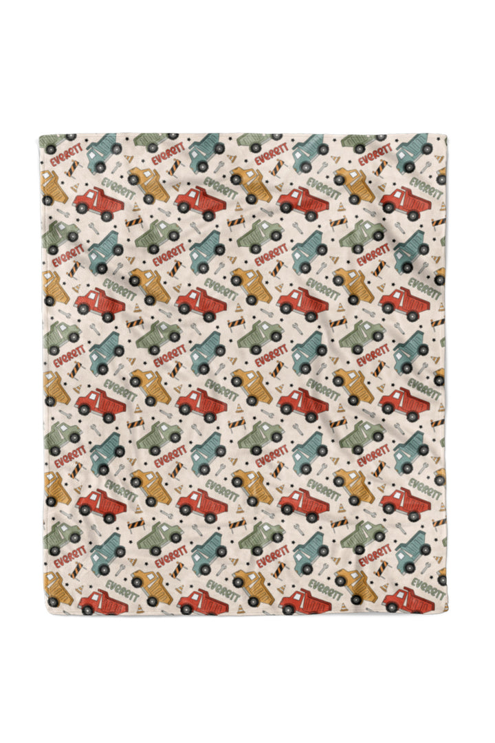 Flat lay of a Caden Lane cream plush blanket in a Busy Builder print featuring red, mustard, sage, and blue dump trucks, reading "Everett" in alternating red and green.
