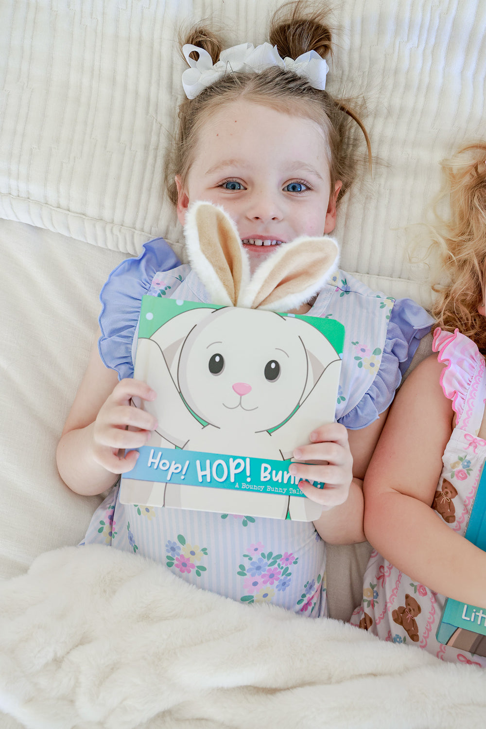 Young girl with bunny ear headband holding Hop HOP Bunny Cuddle Plush Friends Story Book while lying on a bed
