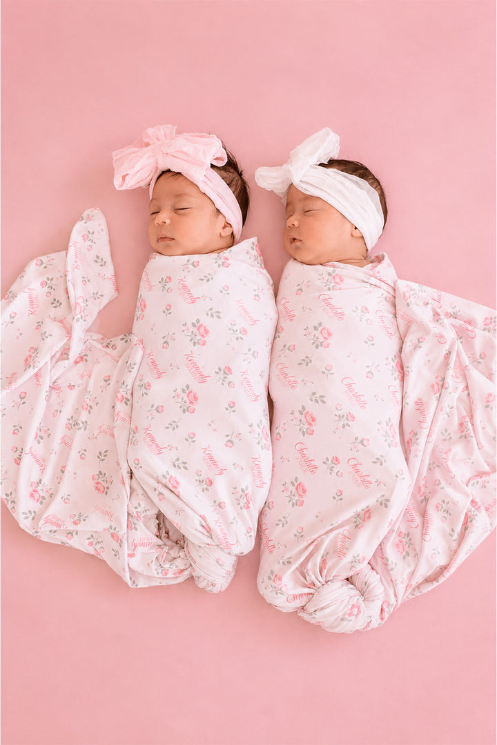Caden Lane personalized swaddle blankets, twin newborns swaddled in matching Rose Garden pink and white floral swaddles with custom names and bow headbands
