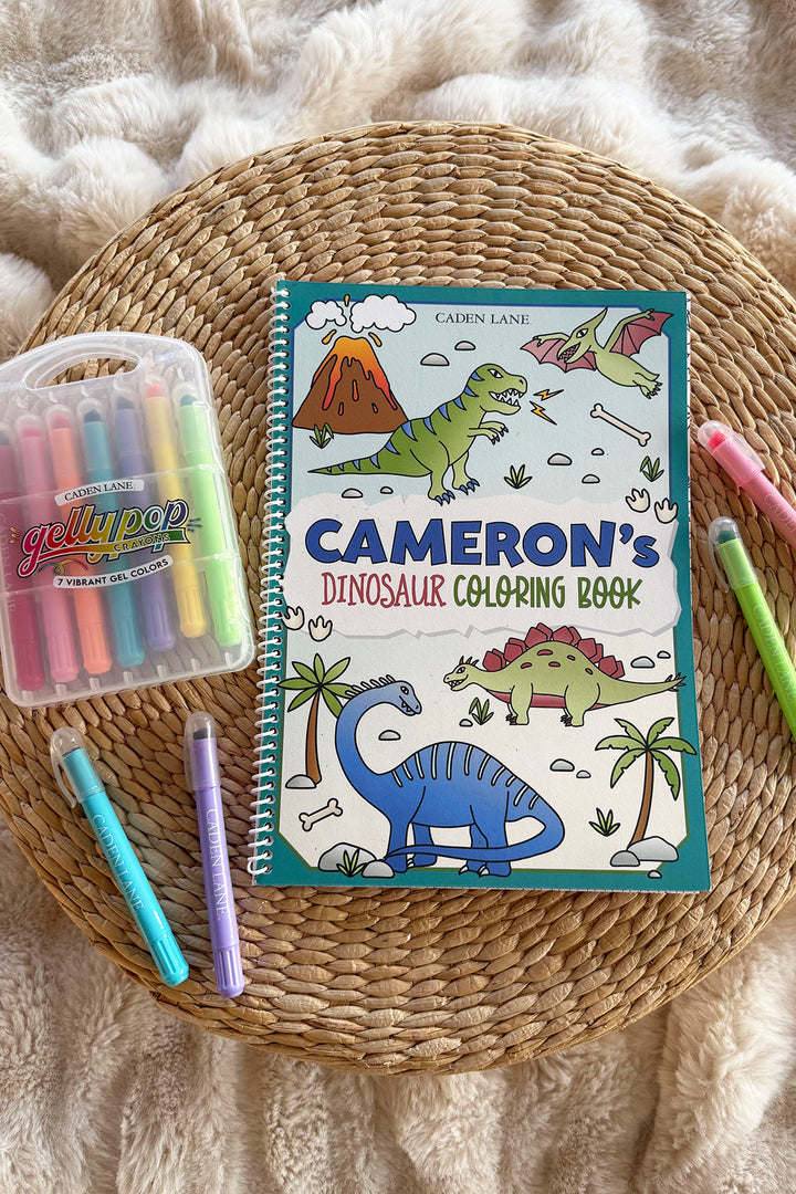 Caden Lane 'Cameron's Dinosaur Coloring Book' on a woven tray with a fur throw, Gellypop gel crayon case, and pastel Caden Lane markers