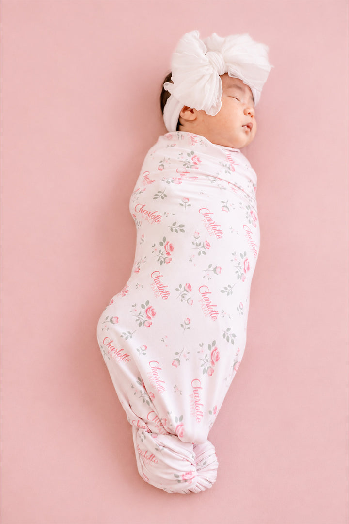Caden Lane personalized newborn swaddle, baby swaddled in Rose Garden pink floral blanket with custom name and white bow headband