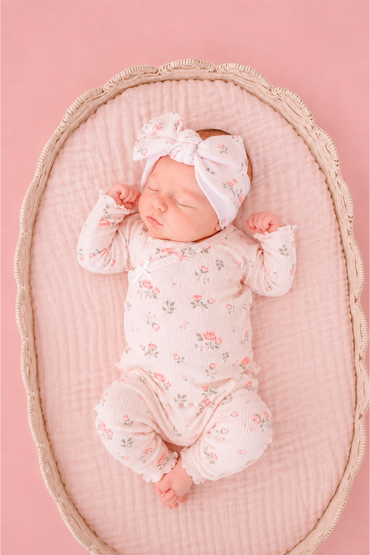 Caden Lane Rose Garden newborn take-home outfit, sleeping baby in bassinet wearing pink floral footie and matching bow headband