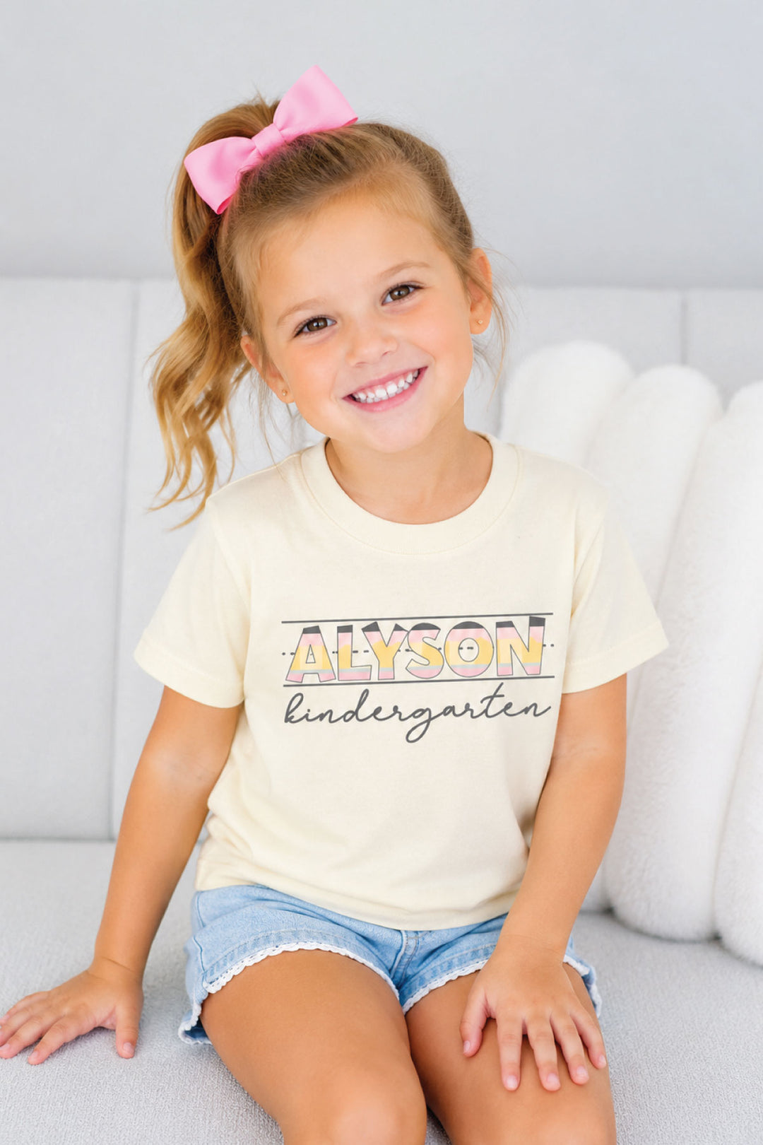 A young girl with a pink hair bow sits on a white sofa wearing a cream-colored Caden Lane t-shirt with 'ALYSON kindergarten' printed on the front in gold and black lettering, paired with denim shorts.