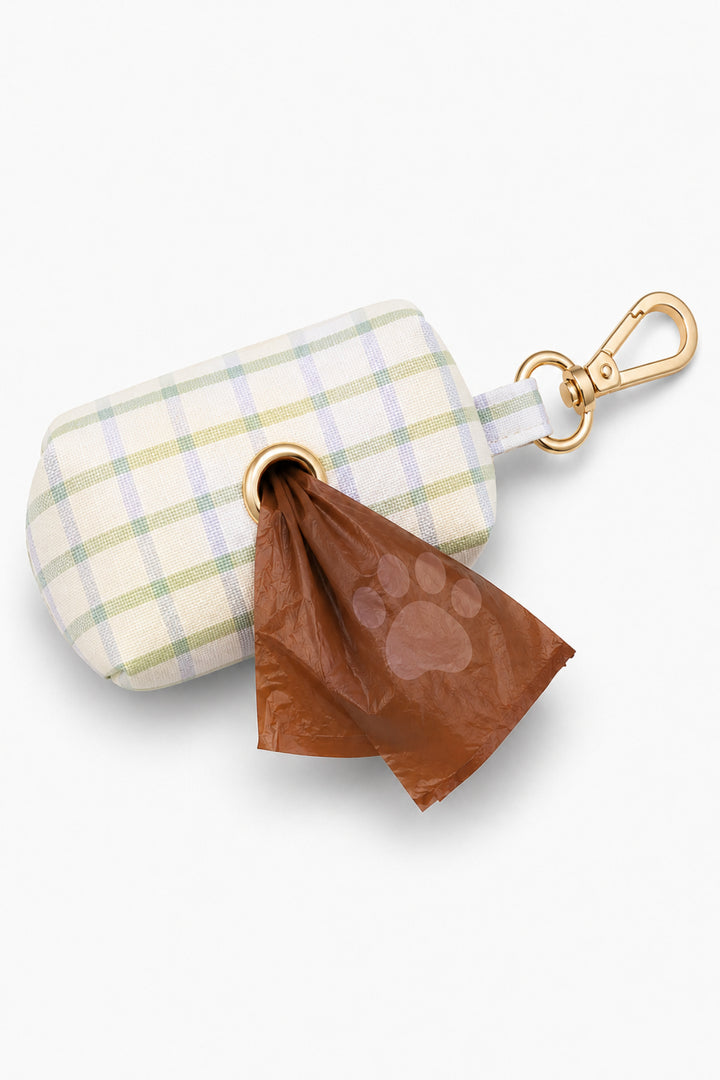 Pet waste bag holder in green and blue picnic plaid shown from the side with a brown waste bag dispensed from the bottom opening and gold carabiner clip attached