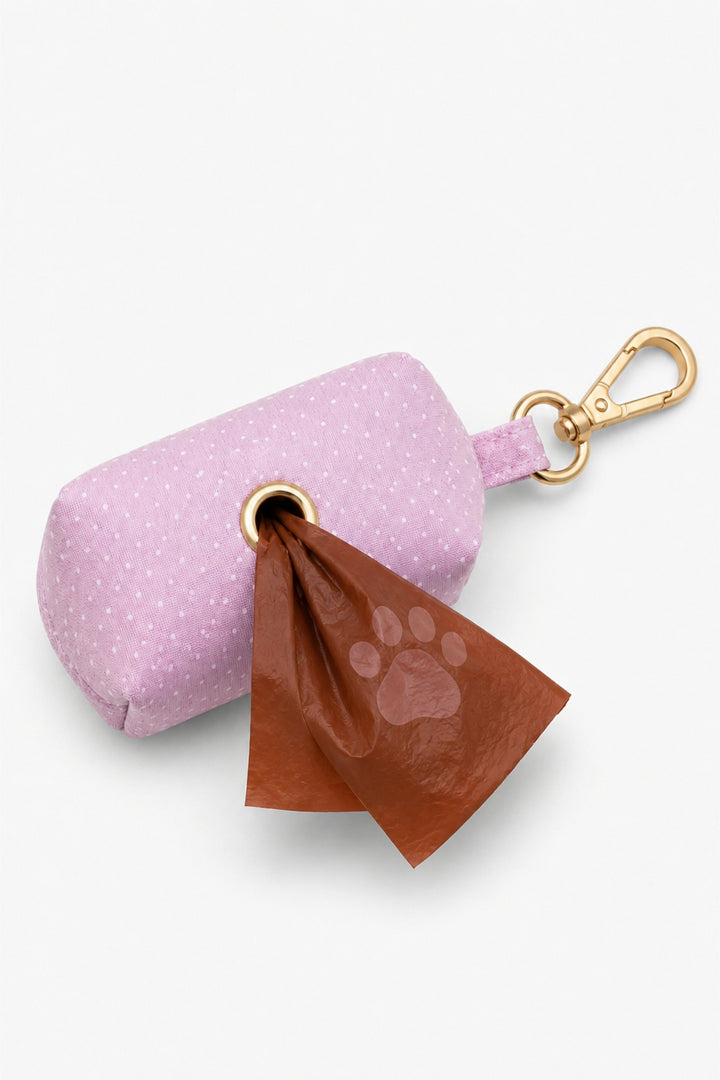 Pink polka dot pet waste bag holder with a brown paw print waste bag being dispensed through the bottom opening and gold carabiner clip