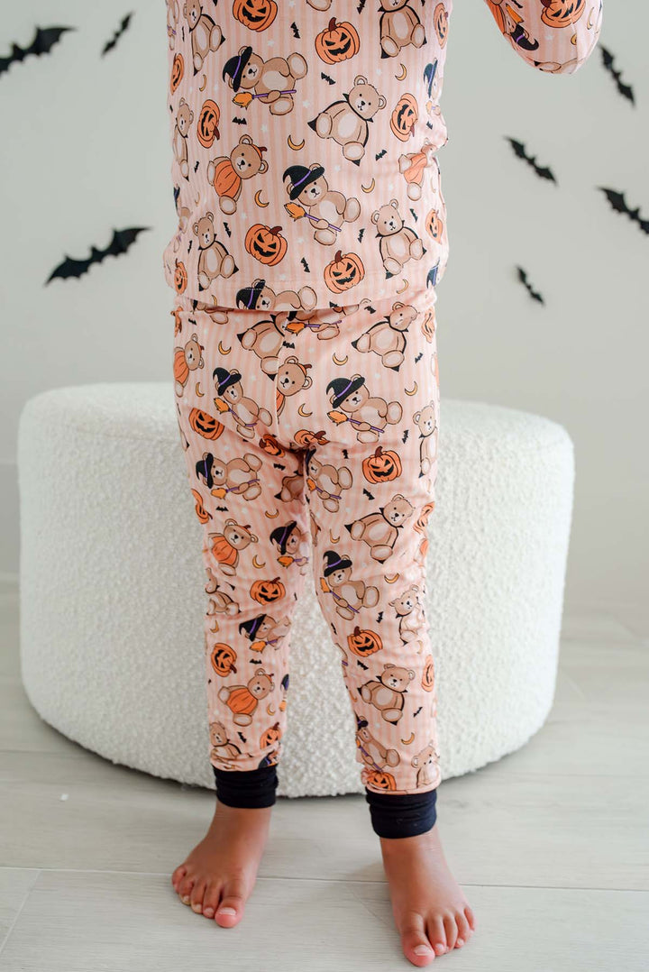 Caden Lane child wearing an orange Halloween-themed pajama set with a teddy bear and pumpkin print, standing barefoot in front of a white textured pouf.