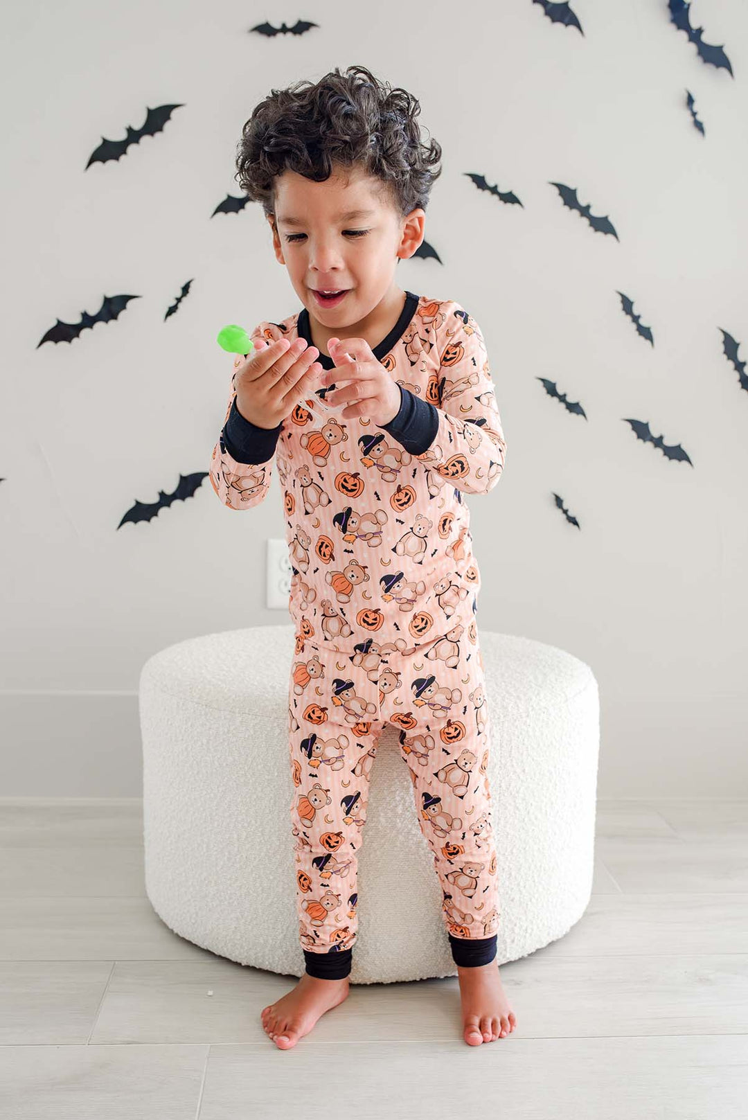Caden Lane young boy wearing a light orange Halloween-themed pajama set with a pumpkin and bat print, sitting on a white round ottoman against a wall decorated with black bat stickers.