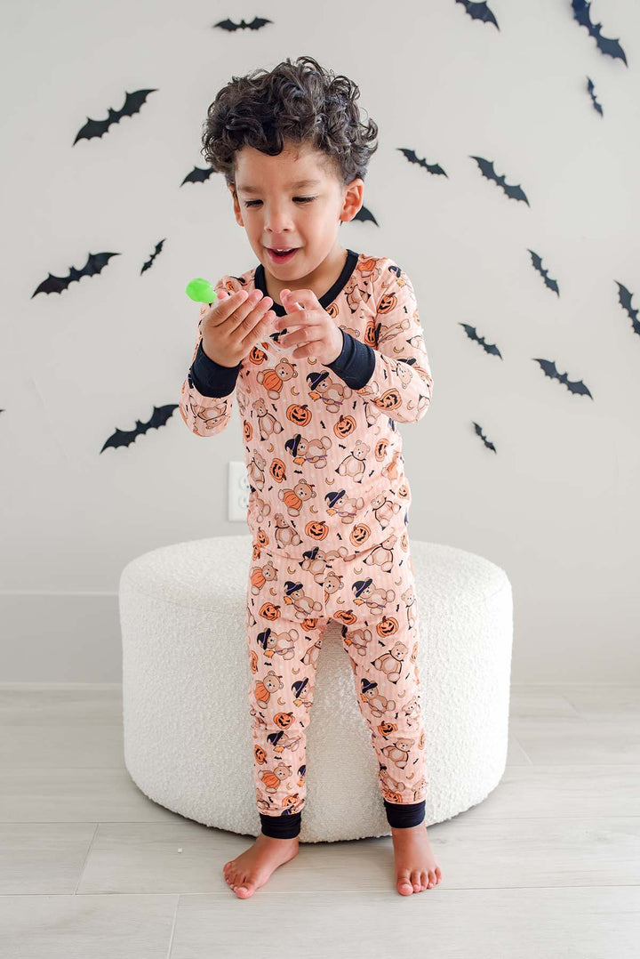 Caden Lane young boy wearing a light orange Halloween-themed pajama set with a pumpkin and bat print, sitting on a white round ottoman against a wall decorated with black bat stickers.