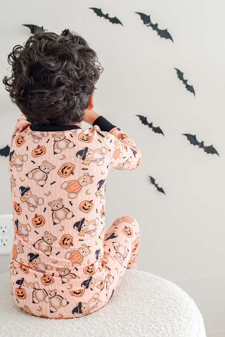 Caden Lane a child seen from behind wearing an orange Halloween-themed pajama set with a print of cartoon cats, pumpkins, bats, and ghosts, sitting on a white textured stool against a white wall decorated with black bat stickers.