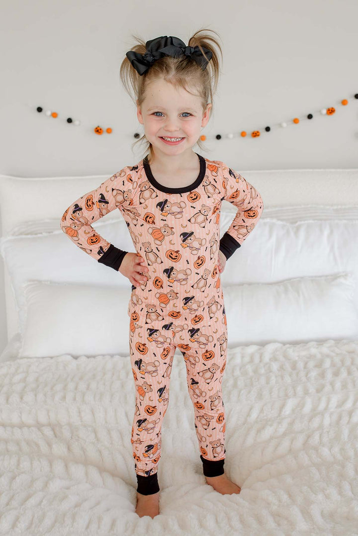 Caden Lane young girl with her hair in two buns wearing an orange Halloween-themed long-sleeve pajama romper with black trim, standing on a white textured bed.