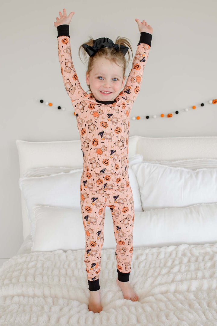 Caden Lane young girl with her hair in a high ponytail wearing a peach-colored Halloween-themed pajama romper, jumping excitedly on a white bed with her arms raised.