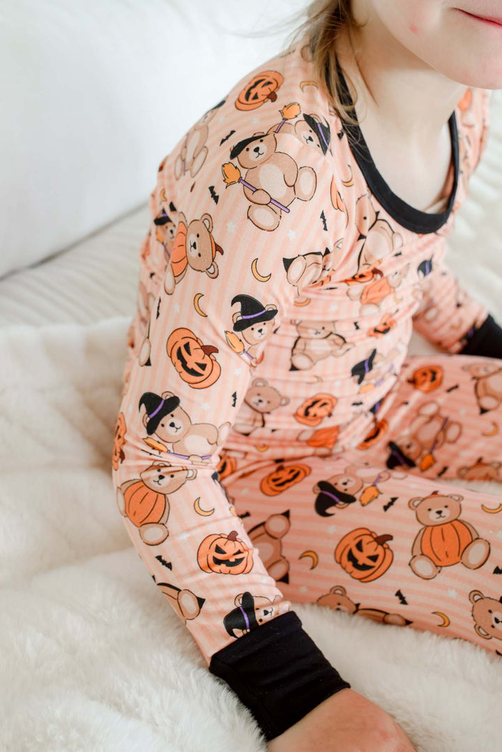 Caden Lane child wearing an orange colored long-sleeve pajama set with a Halloween teddy bear print, sitting on a white bed.