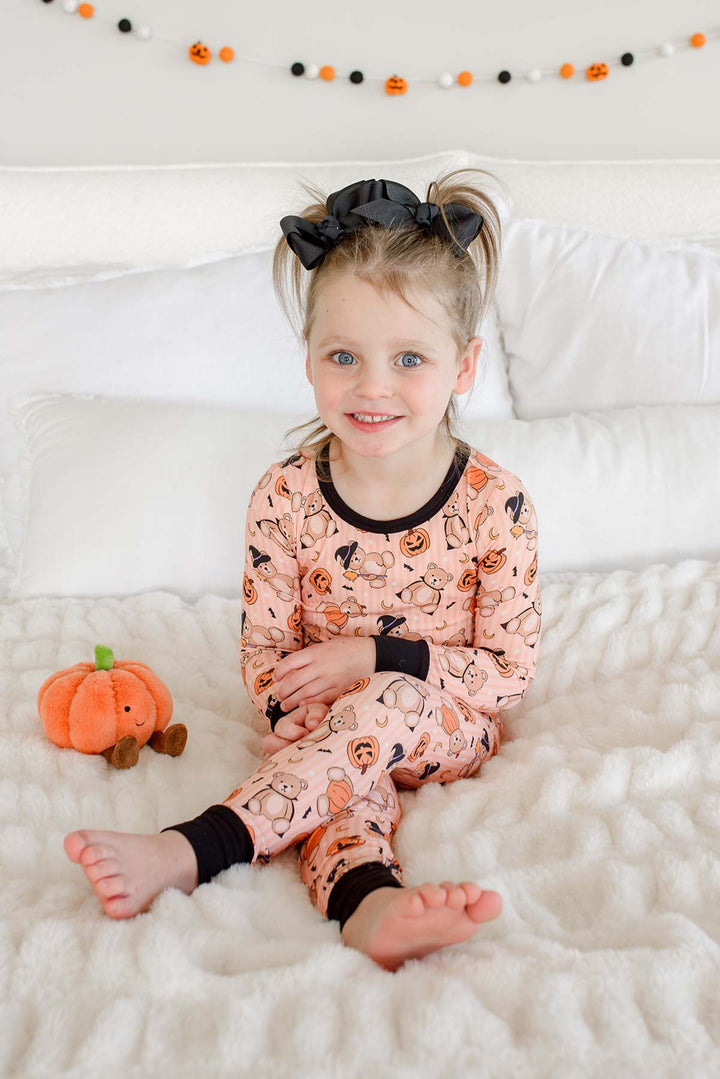 Caden Lane young girl with a black bow in her hair sits on a white bed wearing an orange-colored Halloween pajama set with black trim. A small orange plush pumpkin toy is next to her.
