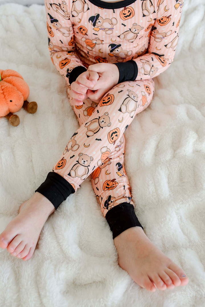 Caden Lane child wearing a light orange long-sleeve pajama set with an all-over Halloween print of pumpkins, ghosts, and bears, sitting on a white textured blanket.