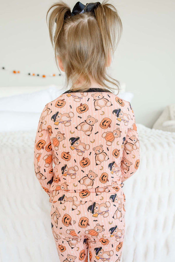 Caden Lane child viewed from behind wearing an orange pajama set with an all-over Halloween print of teddy bears, pumpkins, bats, and ghosts, standing on a white quilted bed.