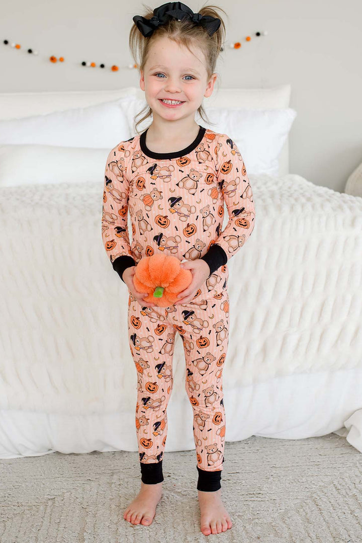 Caden Lane a young girl stands barefoot in a bedroom wearing a long-sleeve and matching pajama set with an orange pumpkin and spider print on a peach background. She is holding a small orange pumpkin prop.