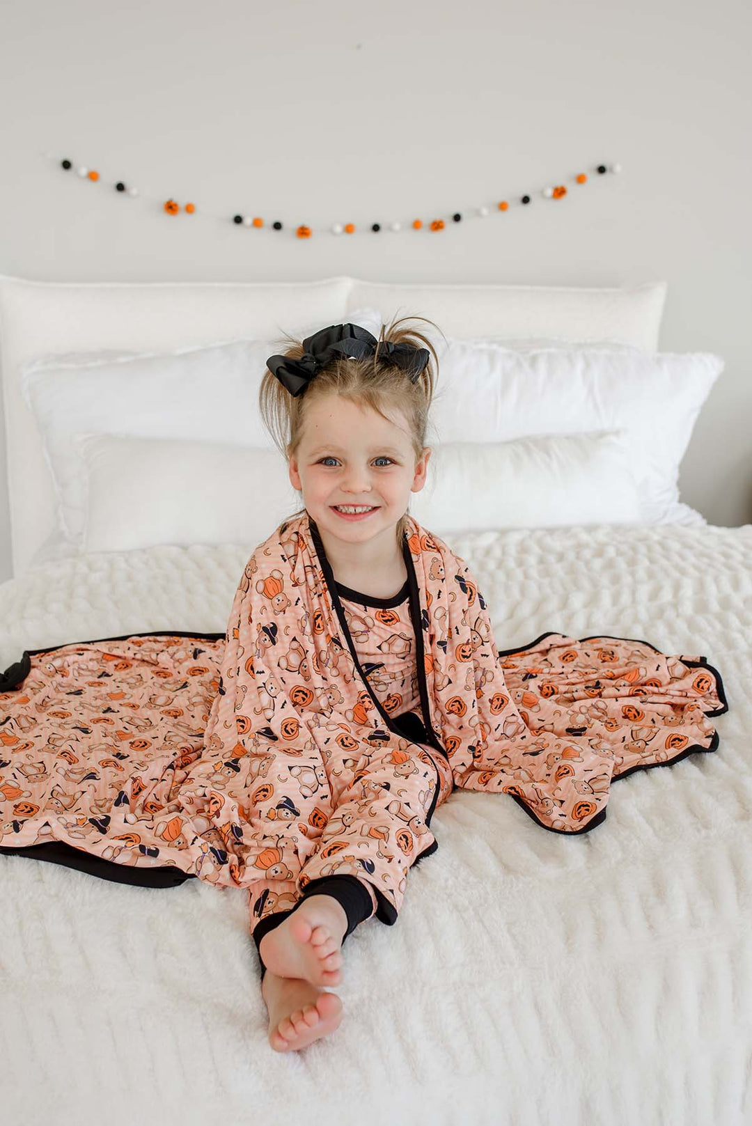 A young girl sitting on a white bed wearing an orange-colored Halloween pajama set with a pumpkin and spider print, along with a matching blanket.