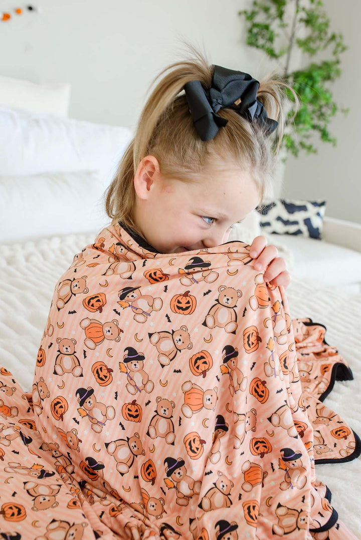 A young girl with blonde hair in a black bow sits on a white bed, wrapped in an orange-colored blanket printed with orange pumpkins, teddy bears, and black witch hats.
