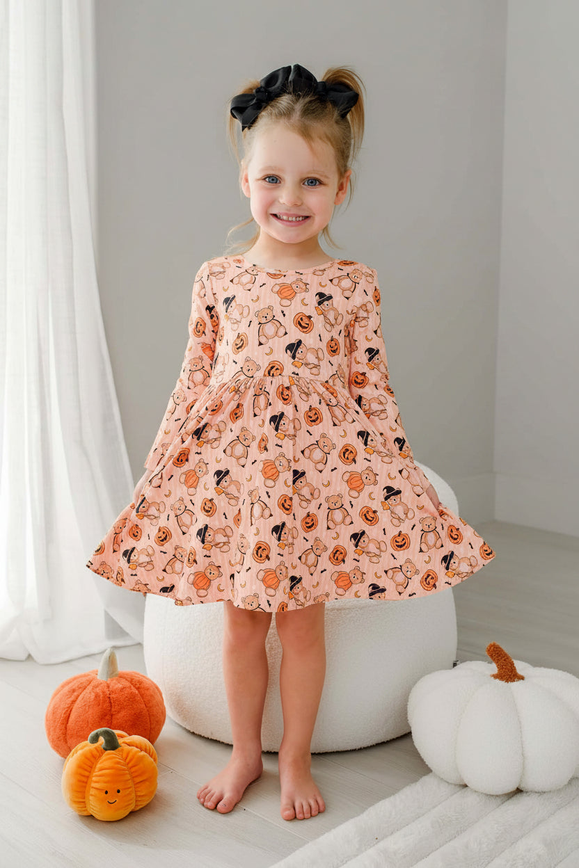 Caden Lane young girl stands barefoot in a bright room wearing an orange long-sleeve dress with an all-over Halloween print. She is posed in front of a white dresser and decorative pumpkins on the floor.