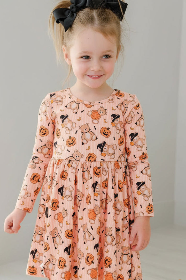 Caden Lane young girl stands in a bright room wearing a long-sleeve orange dress with an all-over black cat print. She has a large black hair bow in her hair and is posed next to decorative pumpkins and a white chair.