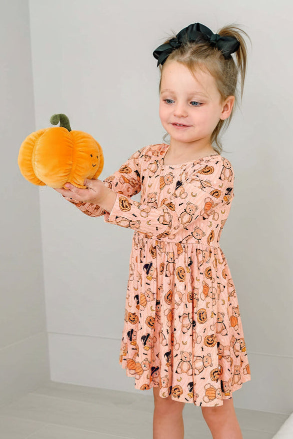Caden Lane young girl holding a plush orange pumpkin. She is wearing an orange-colored long-sleeve dress with a black spider print.