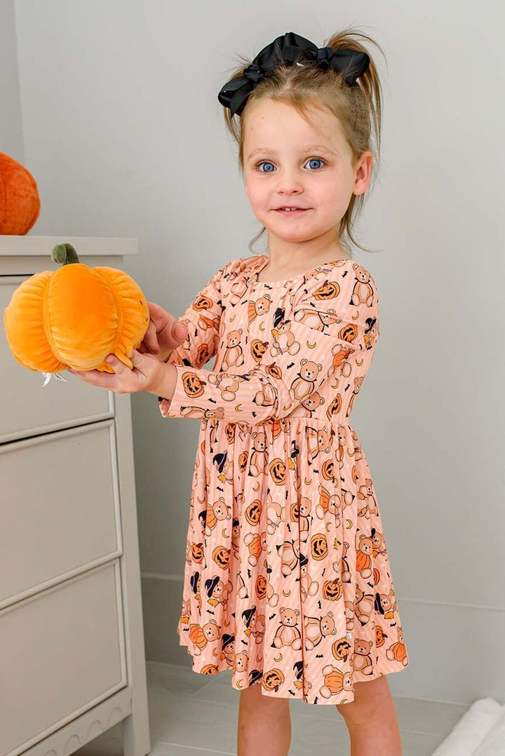 Caden Lane A young girl with a black hair bow stands barefoot in a bedroom, holding a plush orange pumpkin. She is wearing an orange Halloween long-sleeve dress with a black, orange, and white pumpkin print.