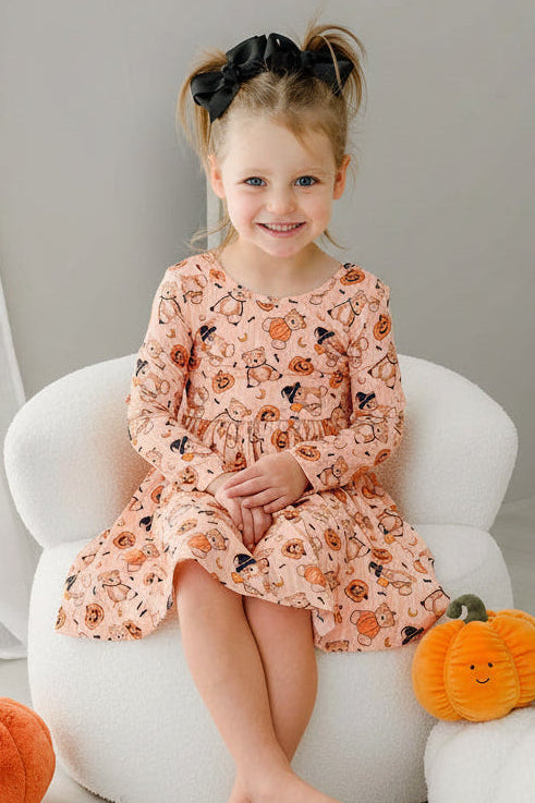 Caden Lane young girl with her hair in two buns sits on a white armchair, wearing an orange long-sleeve dress with an orange and black pumpkin print. The scene is styled with plush pumpkins on a light-colored floor against a neutral gray wall.