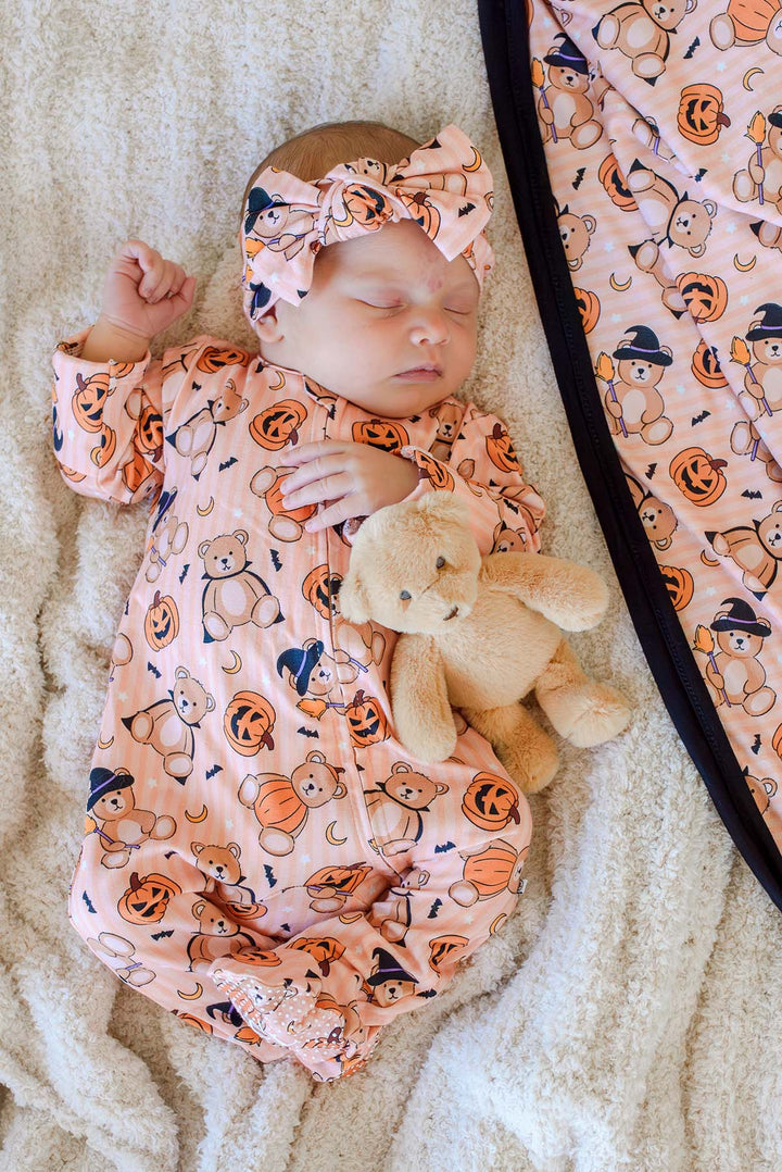 Caden Lane a sleeping newborn baby wearing an orange Halloween-themed footless sleeper and matching bow headband, lying on a cream textured blanket and holding a beige teddy bear. A matching blanket is partially visible on the right.