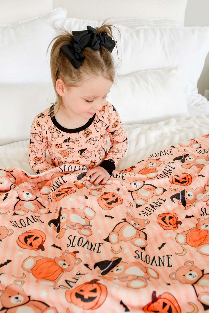 Caden Lane  young girl with a black hair bow sits on a white bed, wearing am orange Halloween-themed long-sleeve pajama top and matching patterned blanket featuring teddy bears, pumpkins, bats, and the name 'Sloane'.