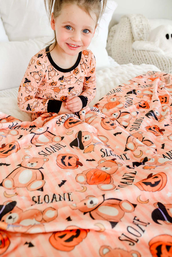 Caden Lane young girl sitting on a bed wearing orange pajamas with a black collar and cuffs, covered by a matching peach fleece blanket printed with orange pumpkins, black bats, and the name 'SLOANE'.
