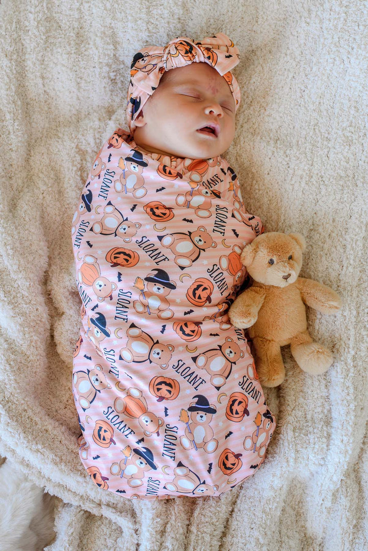 Caden Lane A sleeping newborn baby swaddled in an orange Halloween-themed swaddle blanket and matching headwrap, lying on a soft cream blanket next to a small teddy bear.