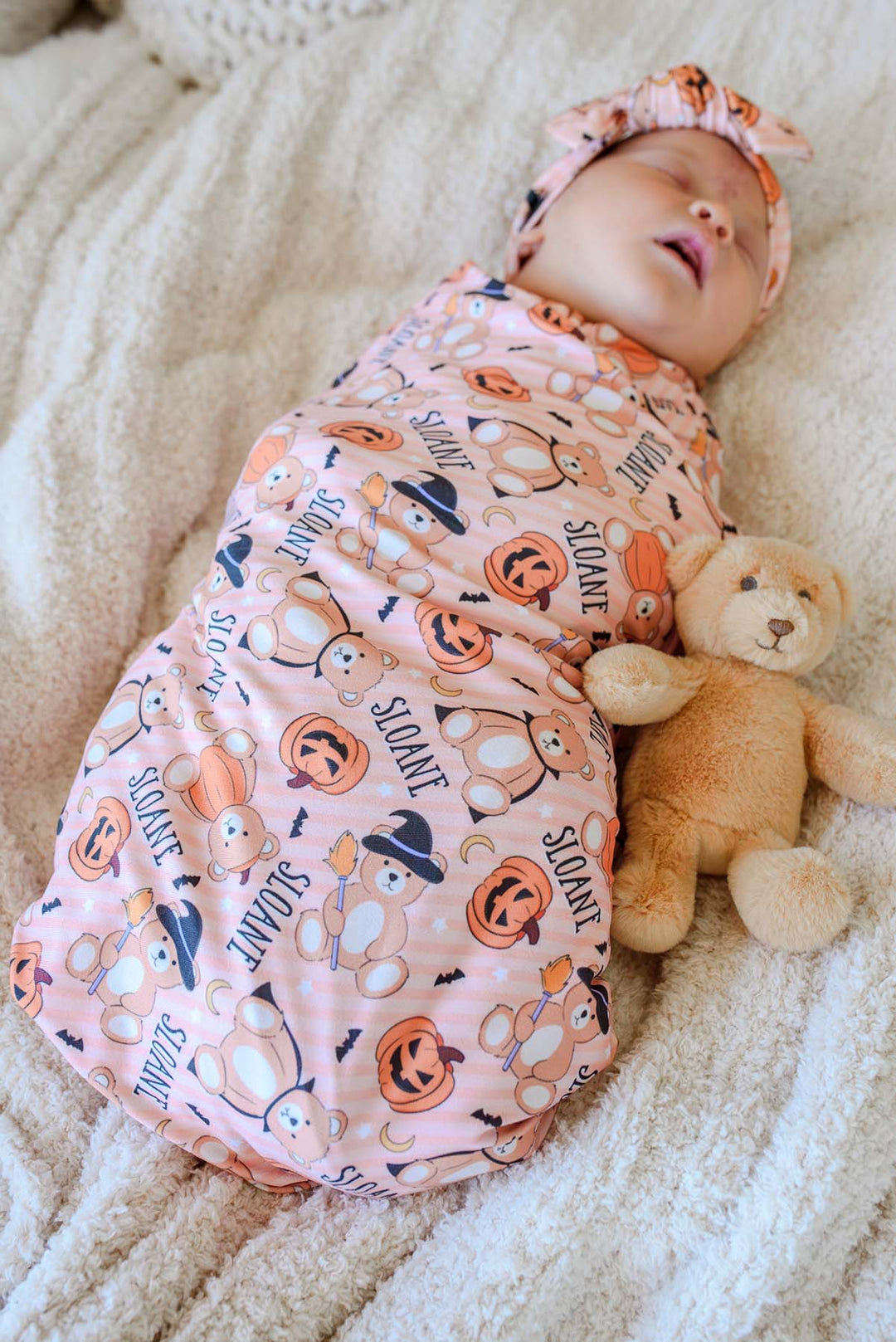 Caden Lane A sleeping newborn baby swaddled in an orange Halloween-themed swaddle blanket with a matching headband, lying on a cream knit blanket next to a teddy bear.
