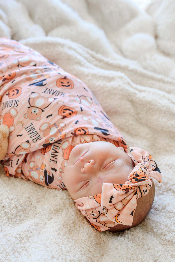 Caden Lane A sleeping newborn baby swaddled in an orange Halloween-themed fabric wrap with orange pumpkins, black bats, and the word 'SNOOE', wearing a matching pink knotted headband, lying on a soft cream-colored blanket.