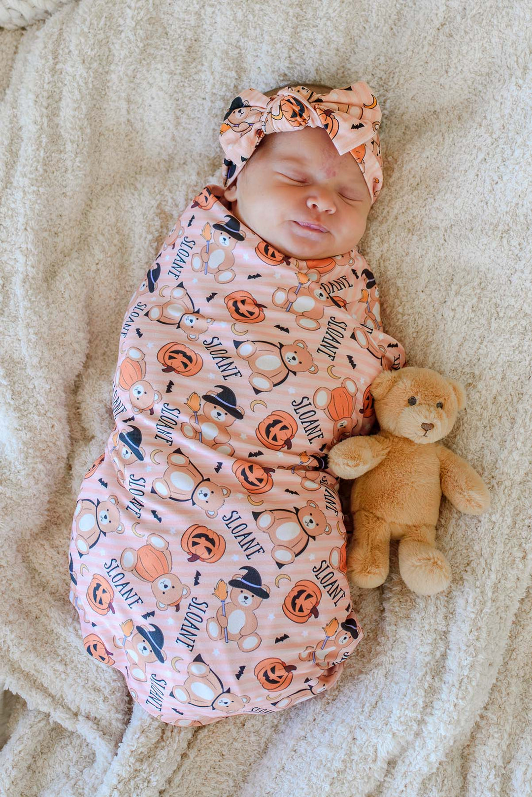 Caden Lane A sleeping newborn baby swaddled in an orange Halloween-themed blanket with a matching headband, lying on a beige blanket next to a small teddy bear.
