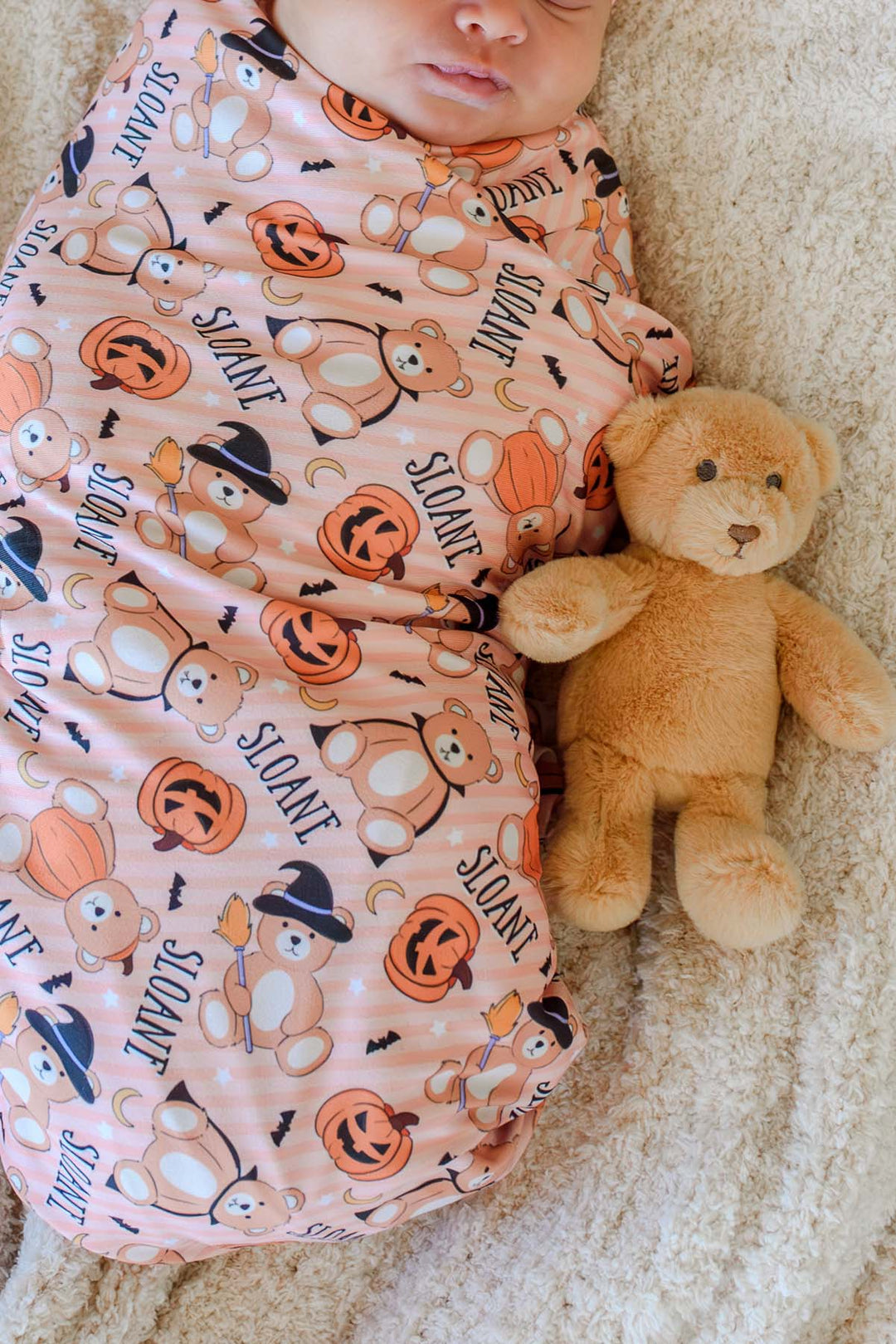 Caden Lane A baby wrapped in an orange Halloween-themed swaddle blanket with a pumpkin, teddy bear, and witch print, lying next to a small brown teddy bear on a textured cream blanket