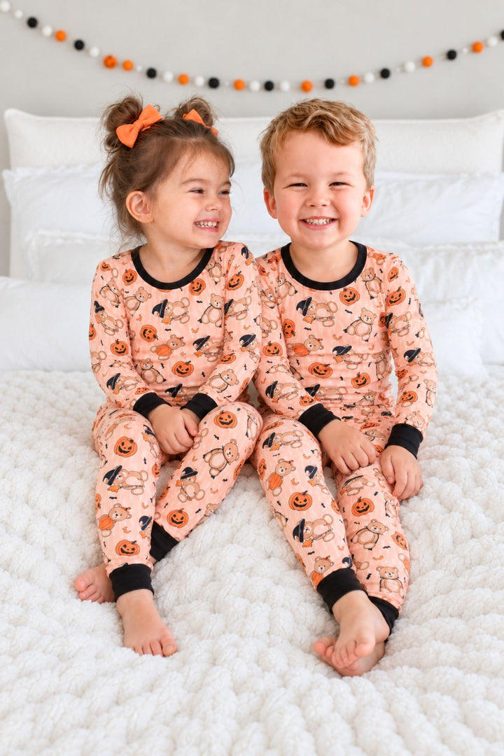 Caden Lane child wearing an orange colored long-sleeve pajama set with a Halloween teddy bear print, sitting on a white bed.