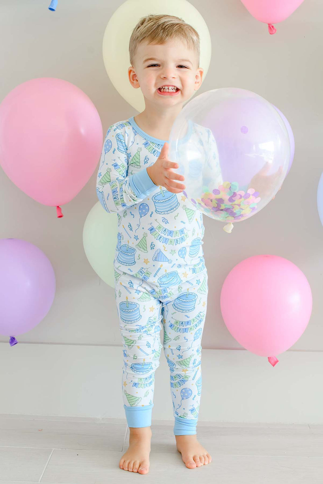 Smiling toddler boy holding a confetti balloon in Caden Lane Birthday Bash Blue birthday pajamas.