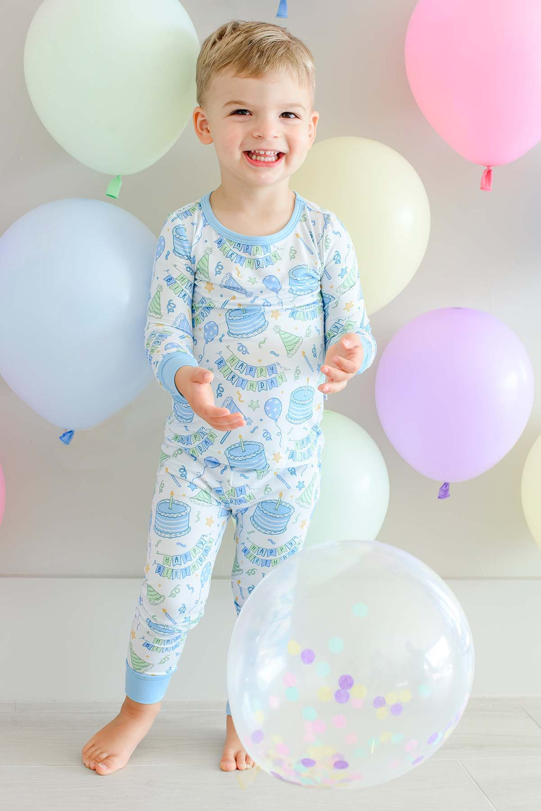 Smiling toddler boy wearing Caden Lane Birthday Bash Blue two-piece pajamas with birthday cakes, balloons and party hats.