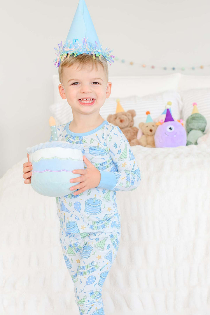 Toddler boy wearing a party hat and Caden Lane Birthday Bash Blue pajamas while holding a plush birthday cake.