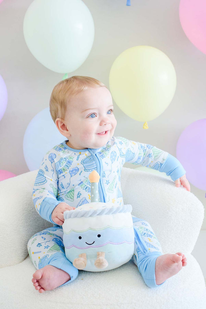 A baby sitting on a white chair wearing a Caden Lane light blue and white patterned footed pajama set, holding a matching plush toy. The background features pastel-colored balloons.
