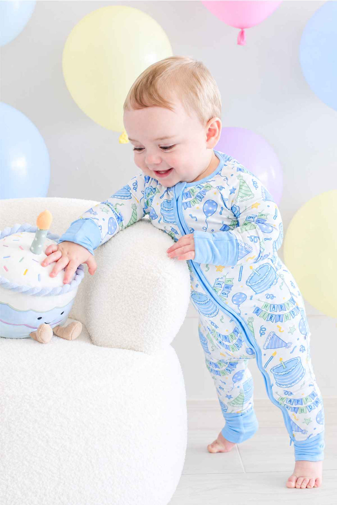 A baby wearing a Caden Lane light blue and white footless one-piece pajama with a colorful balloon print, standing on a white plush chair next to a plush toy against a soft background with pastel balloons.