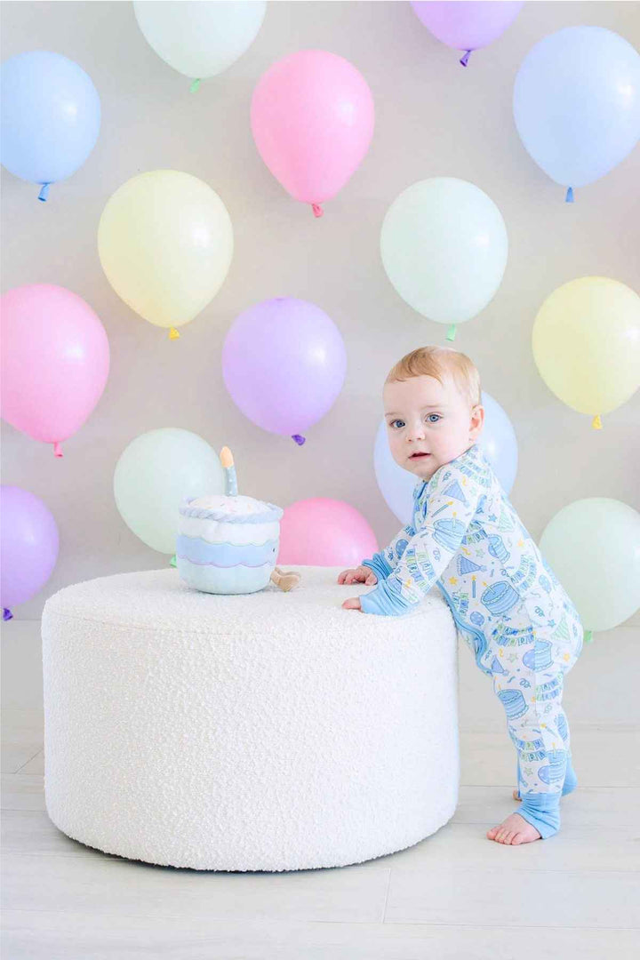 Toddler boy wearing a Caden Lane Birthday Bash zip footie in blue with cake and party print, standing beside a plush birthday cake toy atop a round white ottoman, surrounded by pastel balloons.