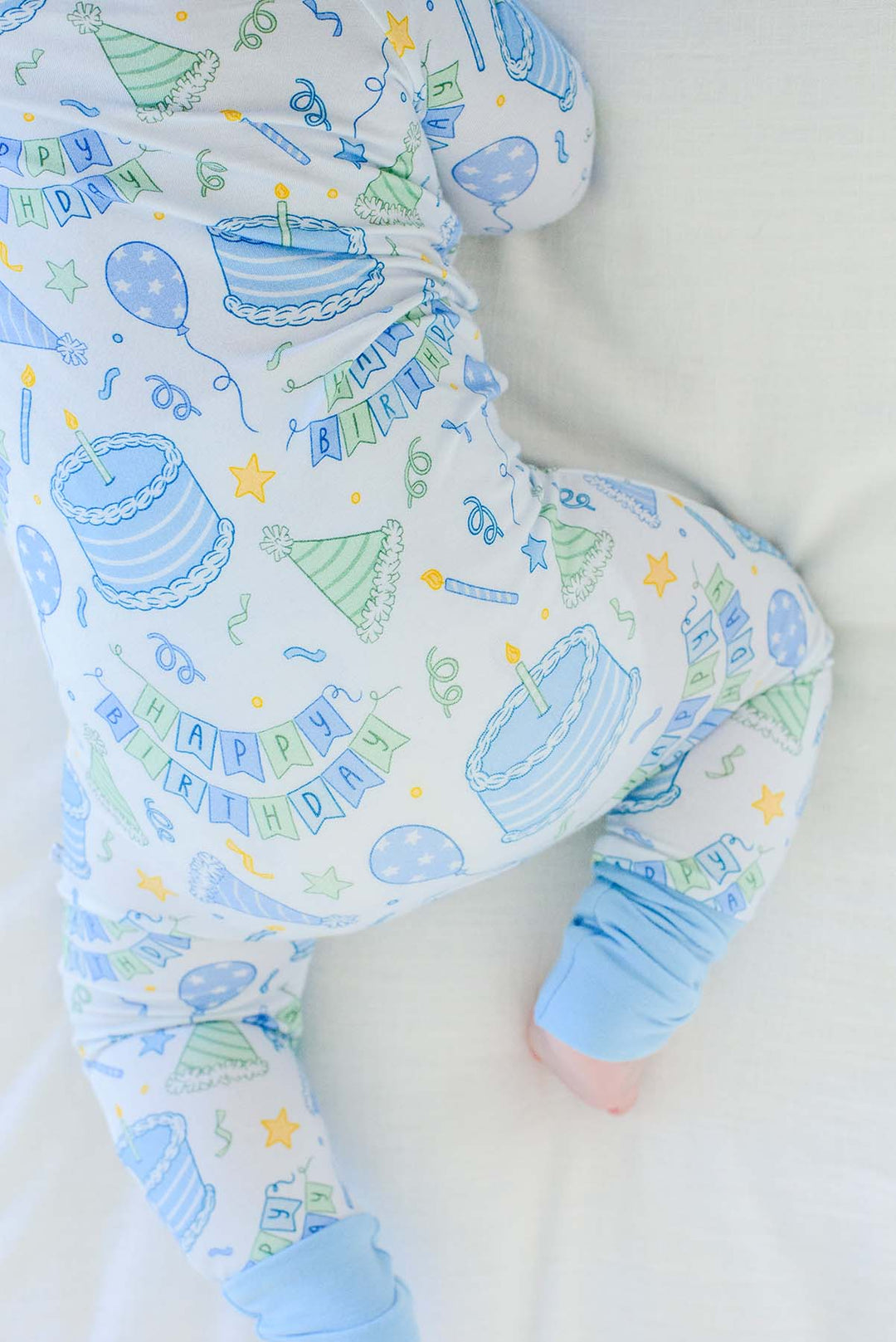 A baby wearing a Caden Lane sleeper with a blue and green birthday party print, lying on a white blanket.