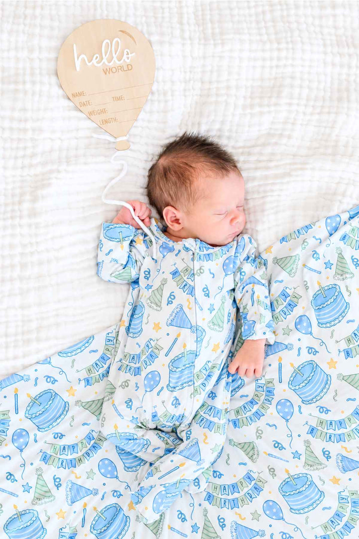 Sleeping newborn wearing a Caden Lane Birthday Bash Blue knotted gown on a matching birthday-print baby blanket.