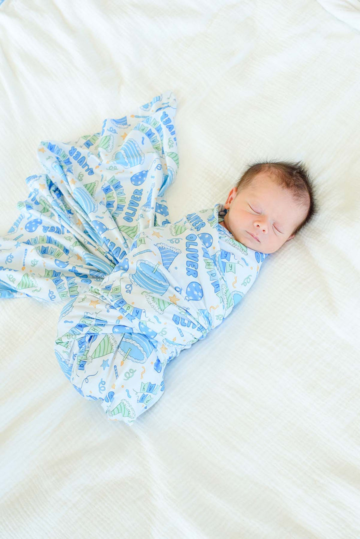 A  baby sleeping in a Caden Lane blue and white patterned swaddle blanket, lying on a white textured bedsheet.