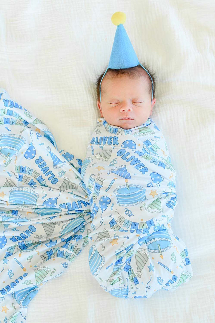 A sleeping baby in a Caden Lane birthday swaddle blanket with a blue and green party print, wearing a blue party hat with a yellow pom-pom. 
